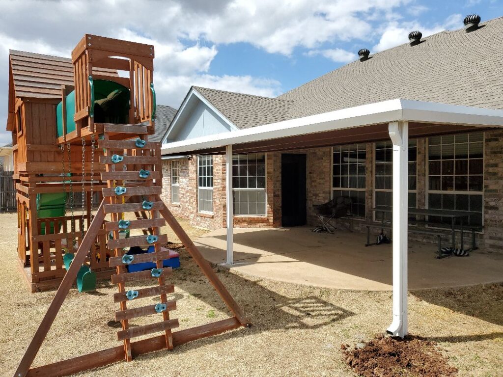 A white patio cover designed by Titan Building Co. is situated next to a wooden playground structure. The cover is attached to a brick house, providing shade to a patio area with a table and chairs.